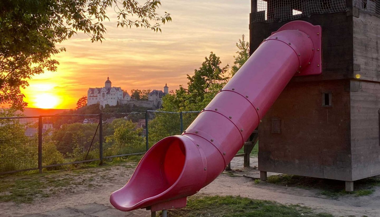 Spielplatz mit roter Rutsche bei Sonnenuntergang und Blick auf historische Burg Ranis in idyllischer Landschaft