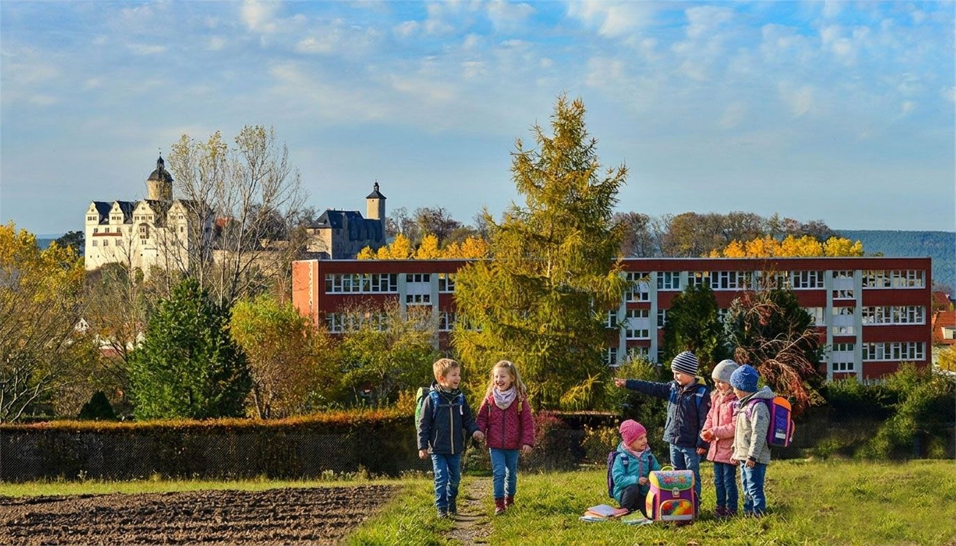 Fröhliche Grundschulkinder mit Schulranzen auf dem Weg zur Schule vor herbstlicher Landschaft und Schulgebäude