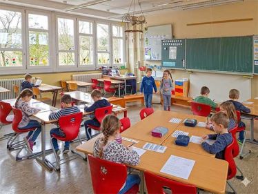 Blick in den Raum mit Tischen, Stühlen und Schultafel und Schülern, die lernen