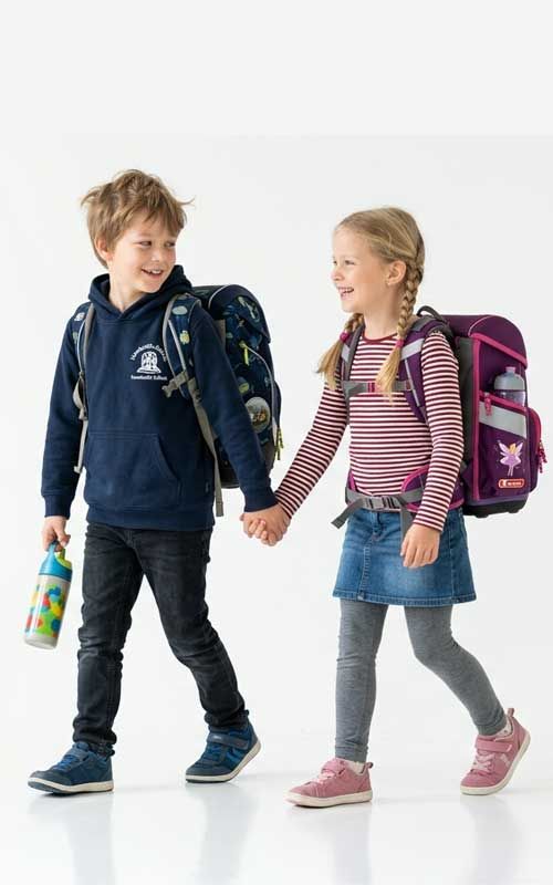 Junge und Mädchen im Grundschulalter mit Schulranzen laufen gemeinsam zur Schule, halten sich an den Händen und wirken fröhlich und entspannt, sie freuen sich auf den Schulstart an der Grundschule Ranis