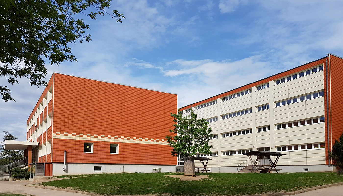 Modernes Schulgebäude der Grundschule mit roter und weißer Fassade, umgeben von Grünflächen und Bäumen unter blauem Himmel