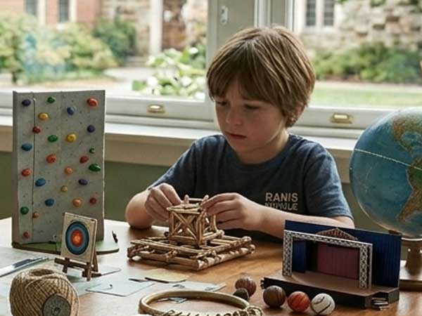 Ein Grundschulkind arbeitet konzentriert an einem handwerklichen Projekt und baut eine geometrische Struktur aus Holzstäbchen. Auf dem Tisch liegen verschiedene Lernmaterialien wie ein Globus, naturwissenschaftliche Modelle und Bastelutensilien.